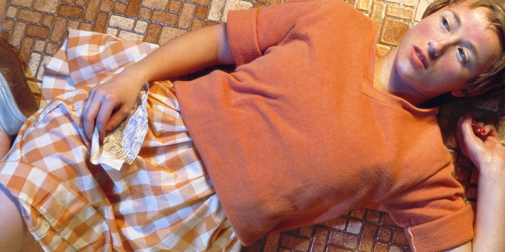 A woman wearing an orange sweater and orange checkered skirt, lying down on a mosaic-tile floor